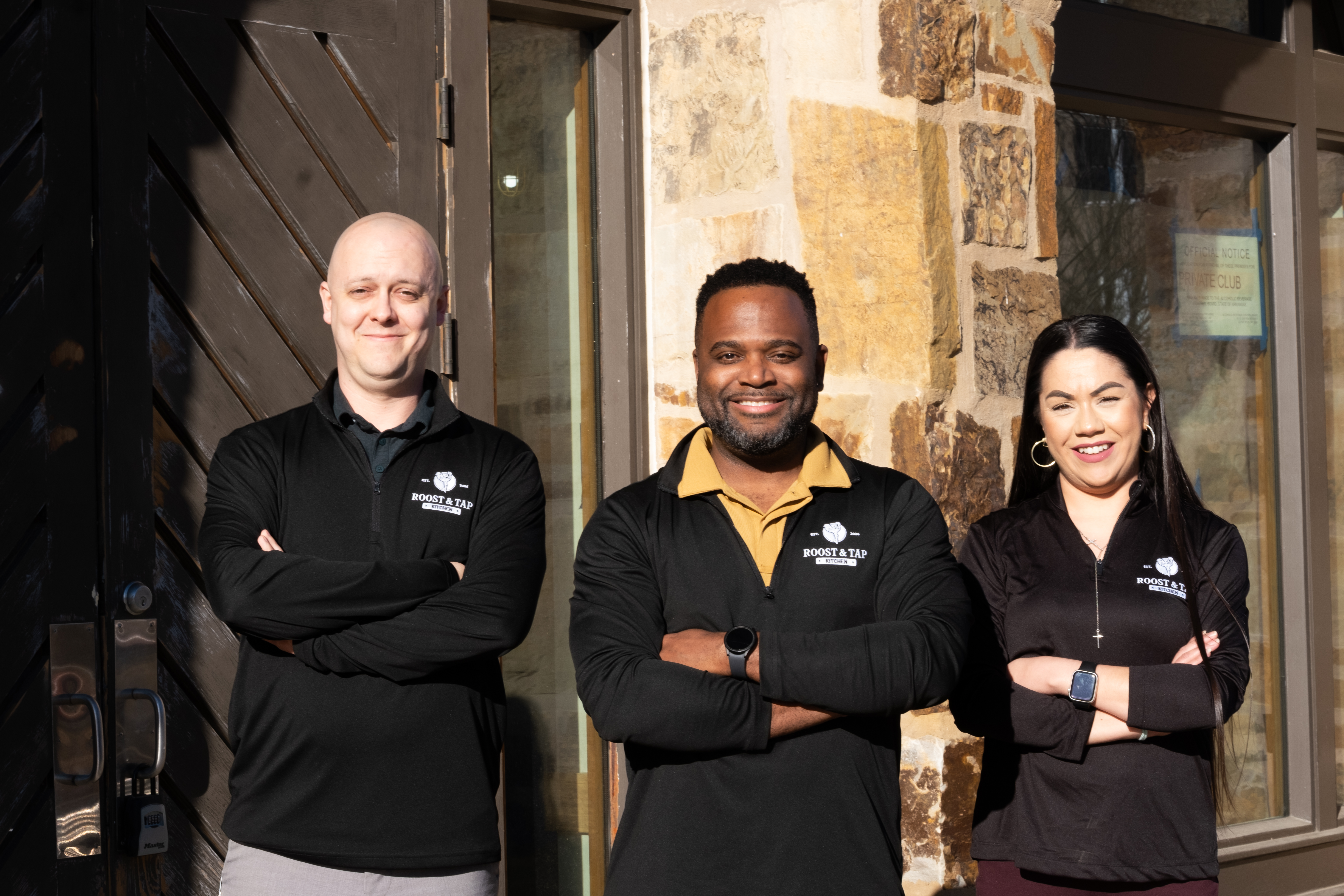 Three staff members standing in front of a stone building, all wearing black jackets with 'Roost & Tap' logo, smiling and posing with arms crossed.