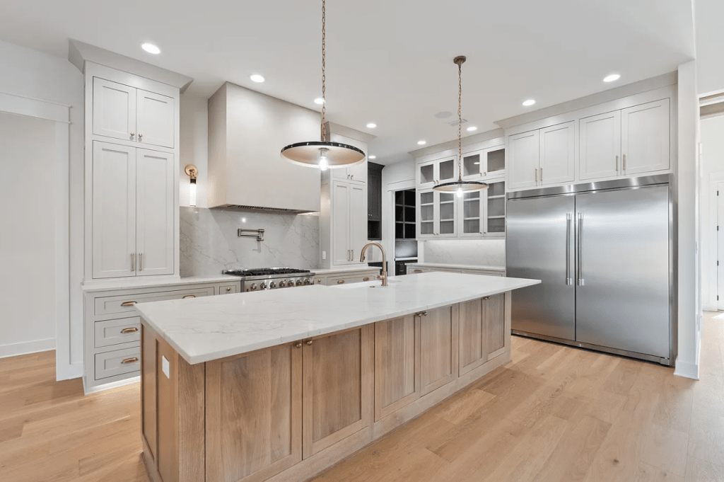 Modern kitchen featuring a large island with a marble countertop, stainless steel appliances, light cabinetry, and warm wood accents.